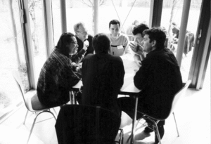 Sechs Männer sitzen in einem hellen Raum an einem Tisch. Einer von ihnen spricht und gestikuliert mit einer Hand.