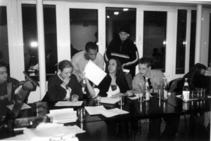 Fünf Jugendliche sitzen an einem Konferenztisch. Zwei Jugendliche beugen sich von hinten über den Tisch und betrachten ein Papier, das eine der Sitzenden ihnen hinhält.