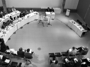 Aus der Vogelperspektive geht der Blick auf ein Konferenzzimmer, wo sich Menschen an weißen Tischen und auf schwarzen Stühlen gegenüber sitzen.