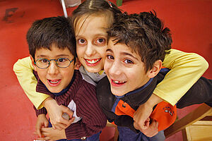 Drei Kinder – ein Mädchen und zwei Jungen im Kindergartenalter – lächeln in die Kamera. Das Mädchen umarmt die Jungen.