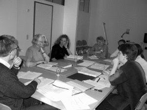 In einem kleineren Raum sitzen an zusammengestellten Tischen fünf Frauen und zwei Männer und tauschen sich aus. Auf den Tischen liegen Papiere und Schreibutensilien.