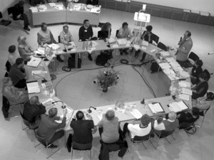 Teilnehmerinnen und Teilnehmer sitzen um einen Tischkreis. Vor ihnen viele Papiere und Mikrofone.