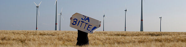Eine Person steht vor einem Windpark und hält ein Schild mit der Aufschrift "JA BITTE!" hoch