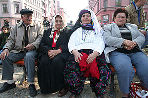 Ein Mann und drei Frauen sitzen an einer Bank auf einem Marktplatz
