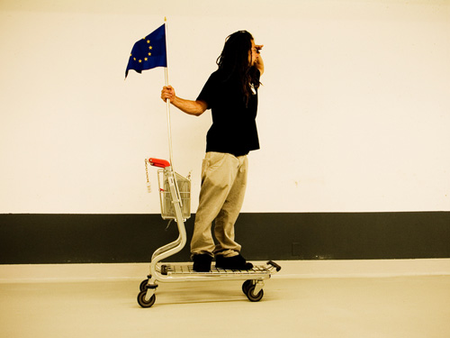 Ein junger Mann steht auf einem Kofferwagen mit einer Europaflagge und blickt in die Ferne
