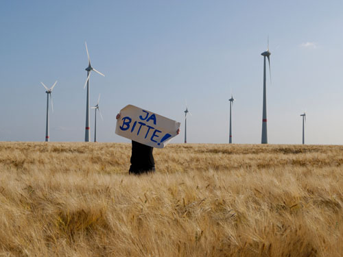 Eine Person steht vor einem Windpark und hält ein Schild mit der Aufschrift "Ja BITTE!" hoch