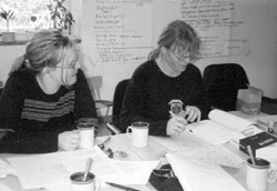 Zwei Frauen sitzen an einem mit Papier bedecktem Tisch. Eine von ihnen notiert etwas auf einem Zettel.